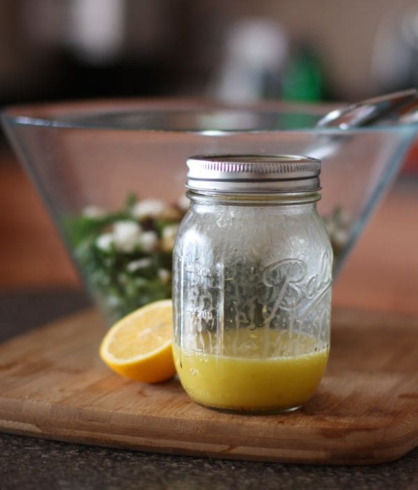mason jar of meyer lemon vinaigrette on cutting board with half of a lemon and bowl of winter kale salad in background