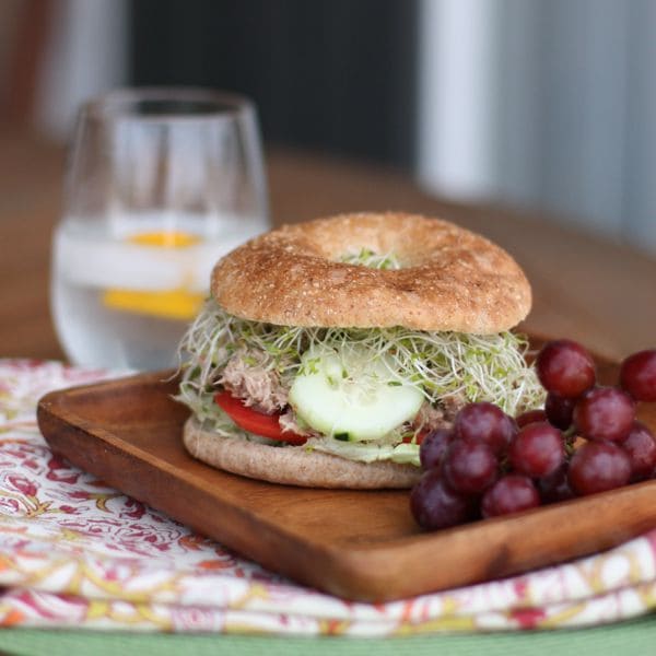 wooden plate with a whole wheat bagel stuffed with tuna salad, sliced tomatoes, lettuce, sliced cucumber, and alfalfa sprouts with a side of red grapes