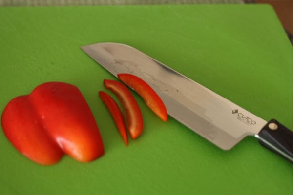 red bell pepper being cut with large knife on a green cutting board