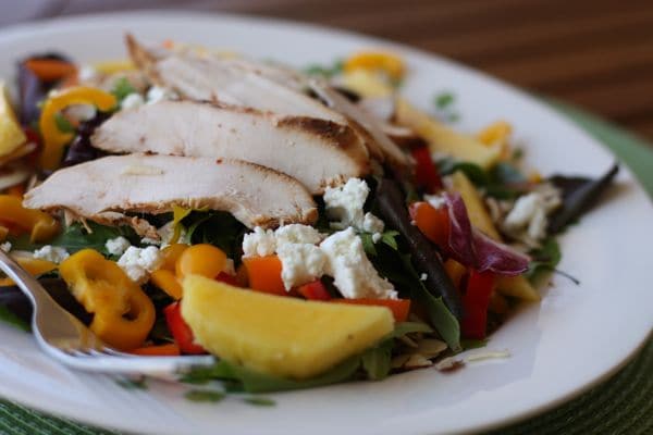 plate of mixed greens topped with yellow and orange bell peppers, sliced mango, goat cheese, almonds, and chicken