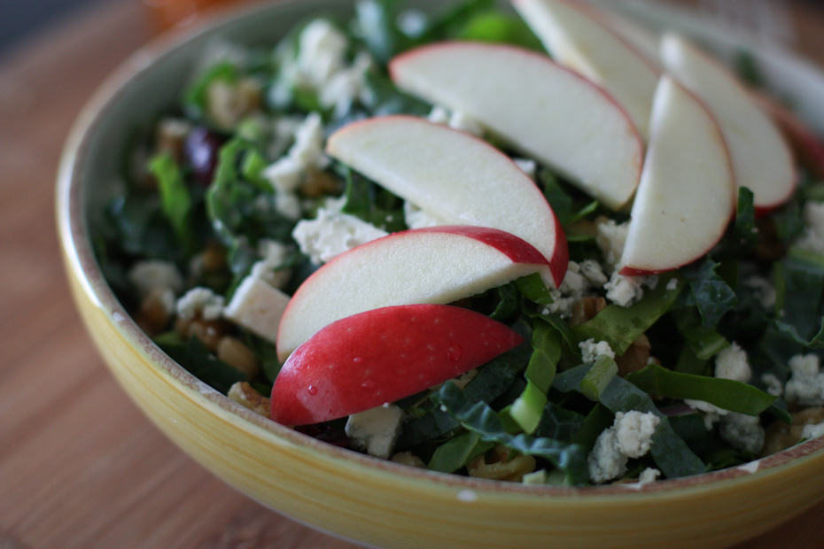 bowl of kale and chard salad topped with apple slices, walnuts, raisins, and bleu cheese crumbles
