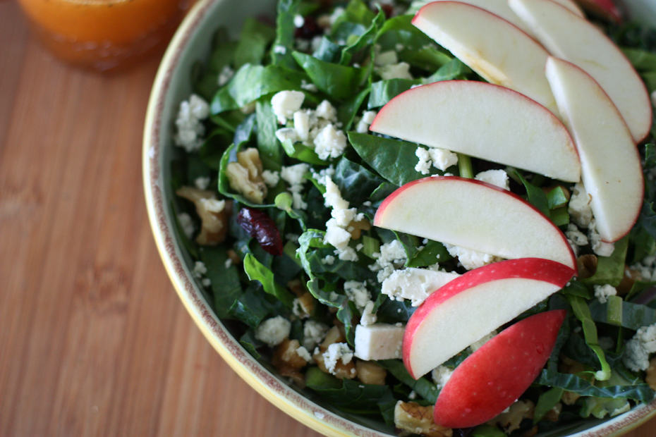 bowl of kale and chard salad topped with apple slices, walnuts, raisins, and bleu cheese crumbles on cutting board