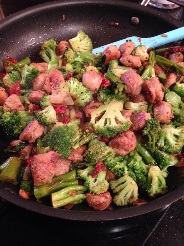 skillet with chicken sausage, broccoli florets, sun dried tomatoes, and onions with blue spatula resting in the skillet 