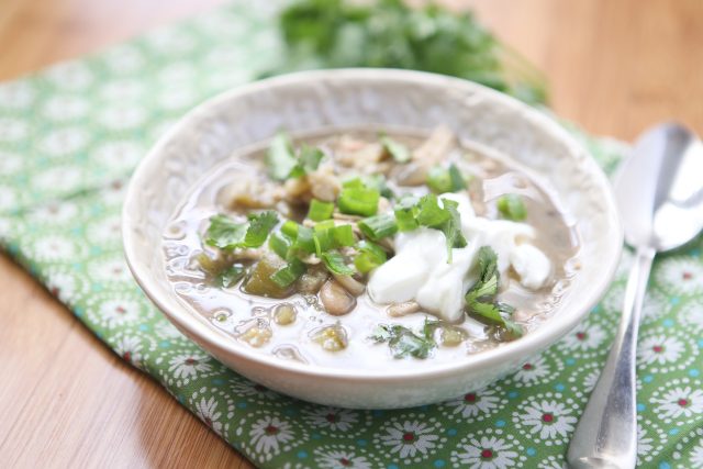 bowl of white chicken chili verde topped with cilantro, green onions, and Greek yogurt on a placemat with a spoon on the side