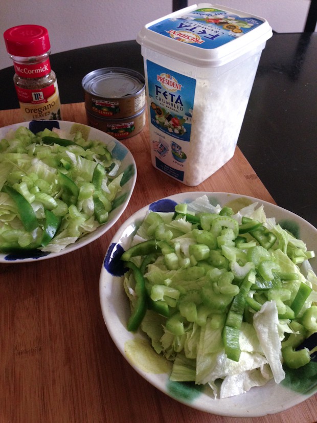 salads being prepared -- two bowls full of lettuce, green bell peppers, and celery alongside oregano leaves and feta
