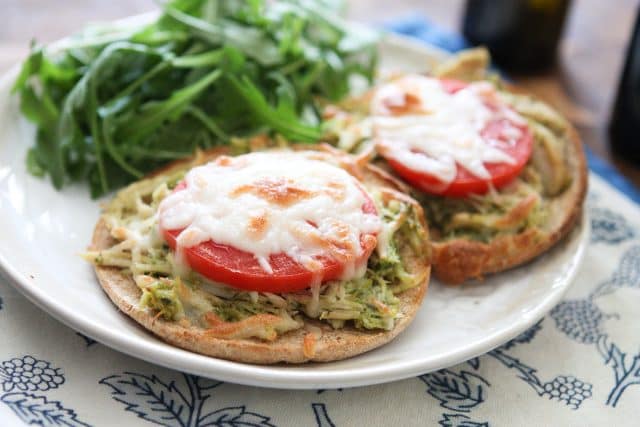 plate with two English muffins topped with shredded chicken, pesto, and tomato with melted mozzarella cheese and a side of arugula