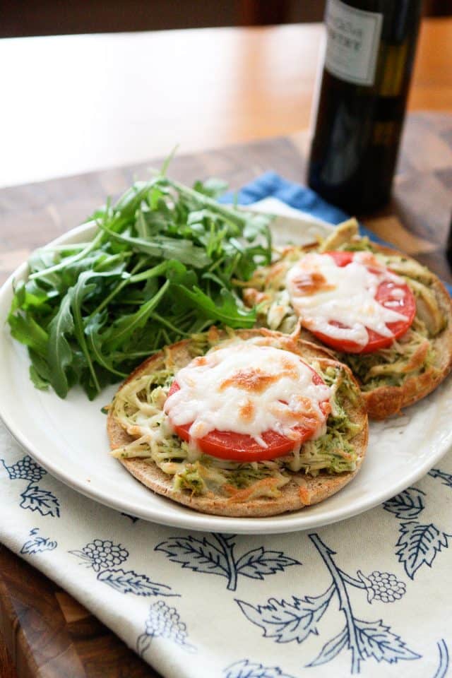 plate with two English muffins topped with shredded chicken, pesto, and tomato with melted mozzarella cheese and a side of arugula