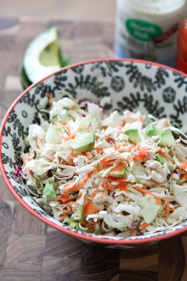 bowl of chicken slaw salad topped with buffalo sauce with a container of blue cheese and half of an avocado in the background