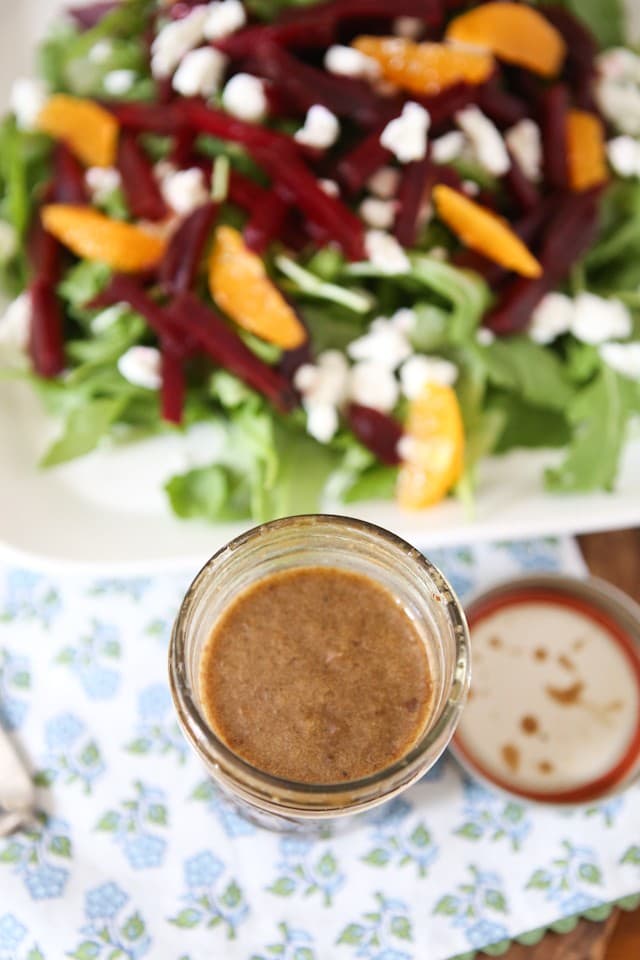 mason jar of dressing next to plate of arugula salad topped with goat cheese, beet strips, orange slices, and walnuts