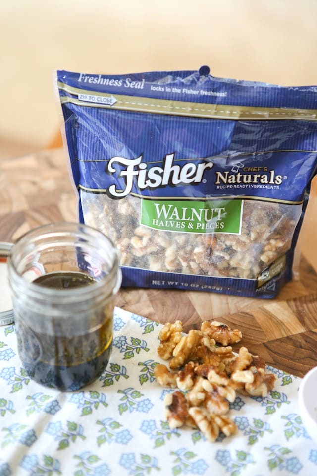 package of Fisher walnut halves and pieces on a cutting board alongside dressing in a mason jar and a handful of the walnuts
