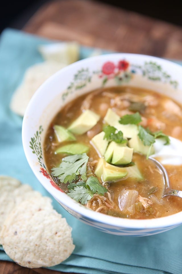 bowl of soup with shredded chicken and various vegetables topped with cilantro and diced avocado with a spoon in the bowl and tortilla chips on the side