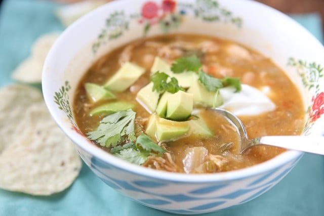 bowl of soup with shredded chicken and various vegetables topped with cilantro and diced avocado with a spoon in the bowl and tortilla chips on the side