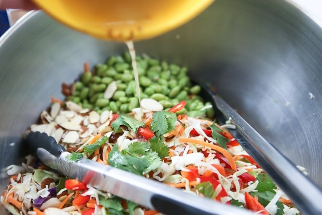 slaw mix, carrots, red bell peppers, edamamae, almond slices, and cilantro in bowl being covered with dressing 