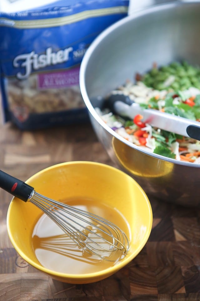 salad dressing being whisked in a small yellow bowl next to the bowl of salad alongside a package of Fisher almond slices