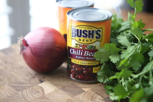 wooden cutting board with a red onion, two cans of Bush's chili bean kidney beans, and bunch of cilantro