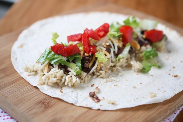 unwrapped tortilla being assembled with brown rice, ground turkey, black beans, shredded cheese, romaine lettuce, and chopped tomatoes