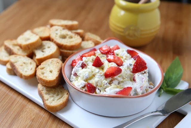 white bowl filled with strawberry basil goat cheese spread on a white platter with crispy toasts for serving