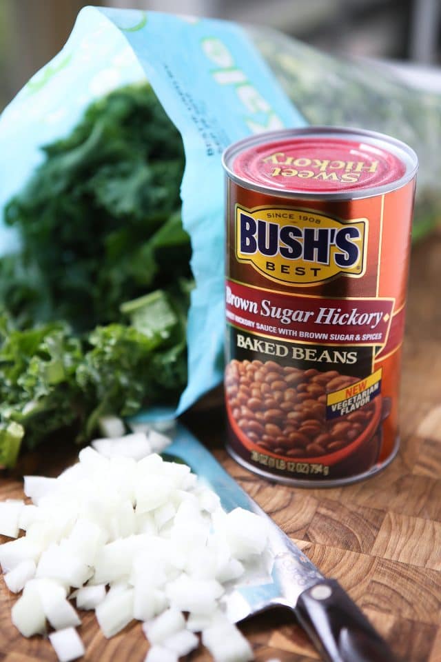 can of Bush's brown sugar hickory baked beans, bunch of kale, and chopped white onions on a cutting board with a knife