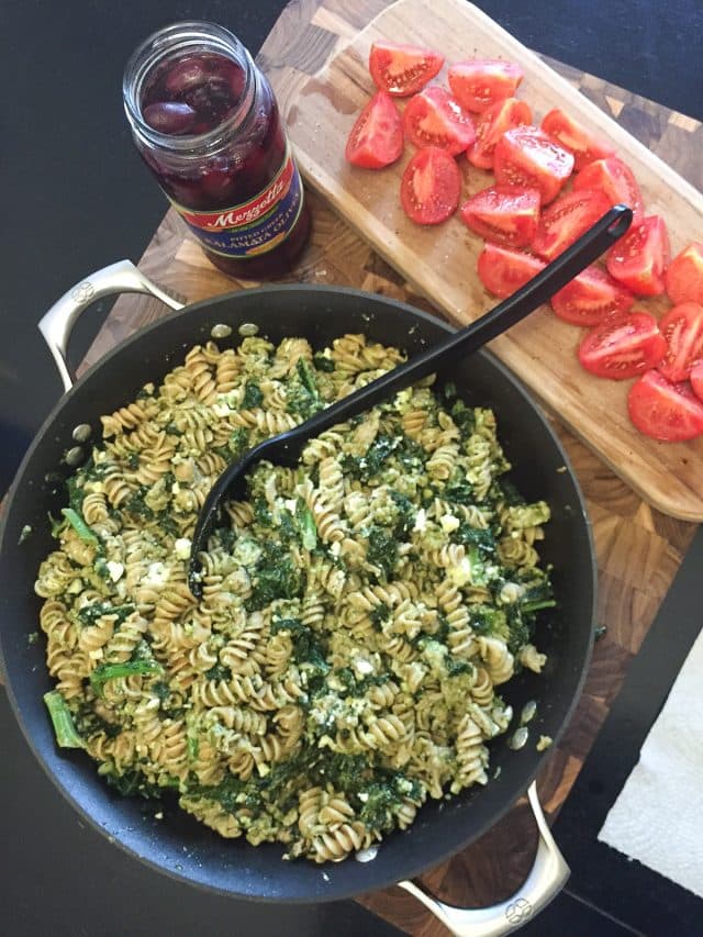 large skillet filled with pesto pasta, ground turkey and kale on a table with quartered tomatoes and greek olives