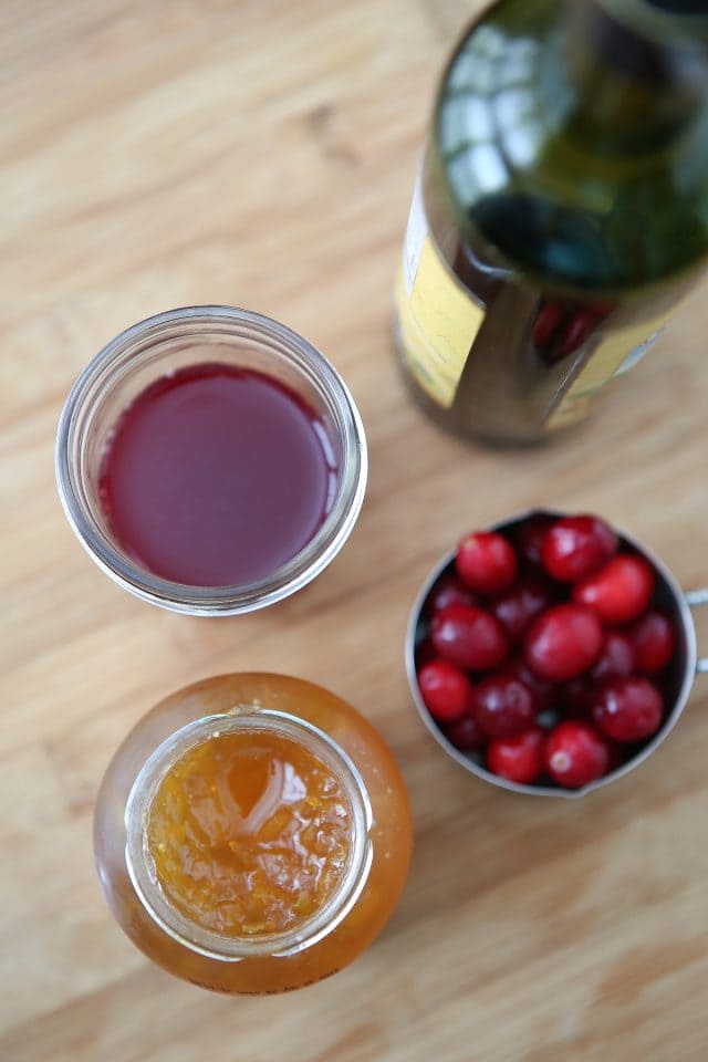 jar of orange marmalade, container of cranberries, and cup of cranberry juice sitting next to its container on a wooden countertop