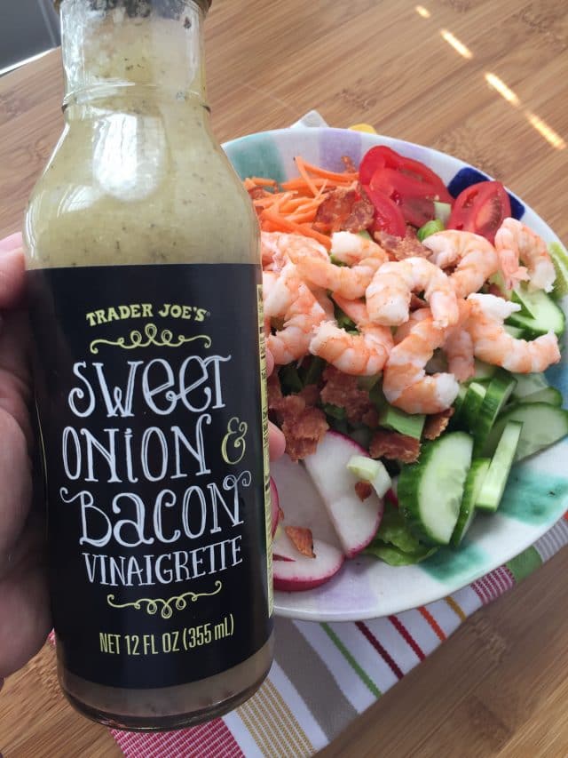 bowl of lettuce topped with cherry tomatoes, shrimp, radishes, cucumbers, carrots, and red peppers with a hand holding Trader Joe's Sweet Onion and Bacon Vinaigrette