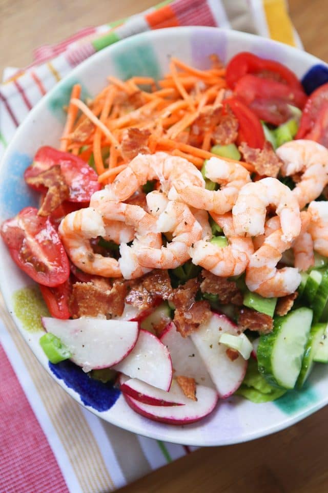 bowl of greens arranged with shrimp, shredded carrots, tomatoes, cucumbers, radishes, and bacon