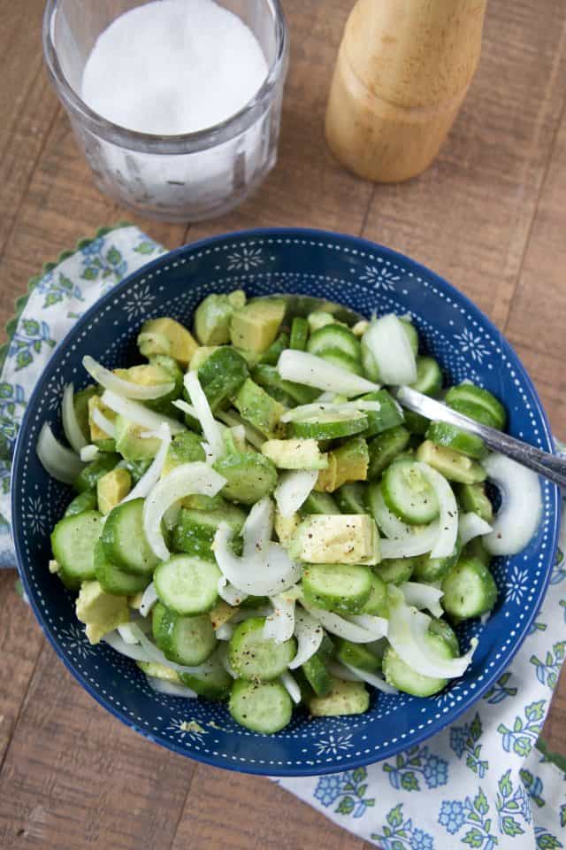 sliced cucumbers, onion and diced avocado in a blue bowl tossed with lemon, salt and pepper